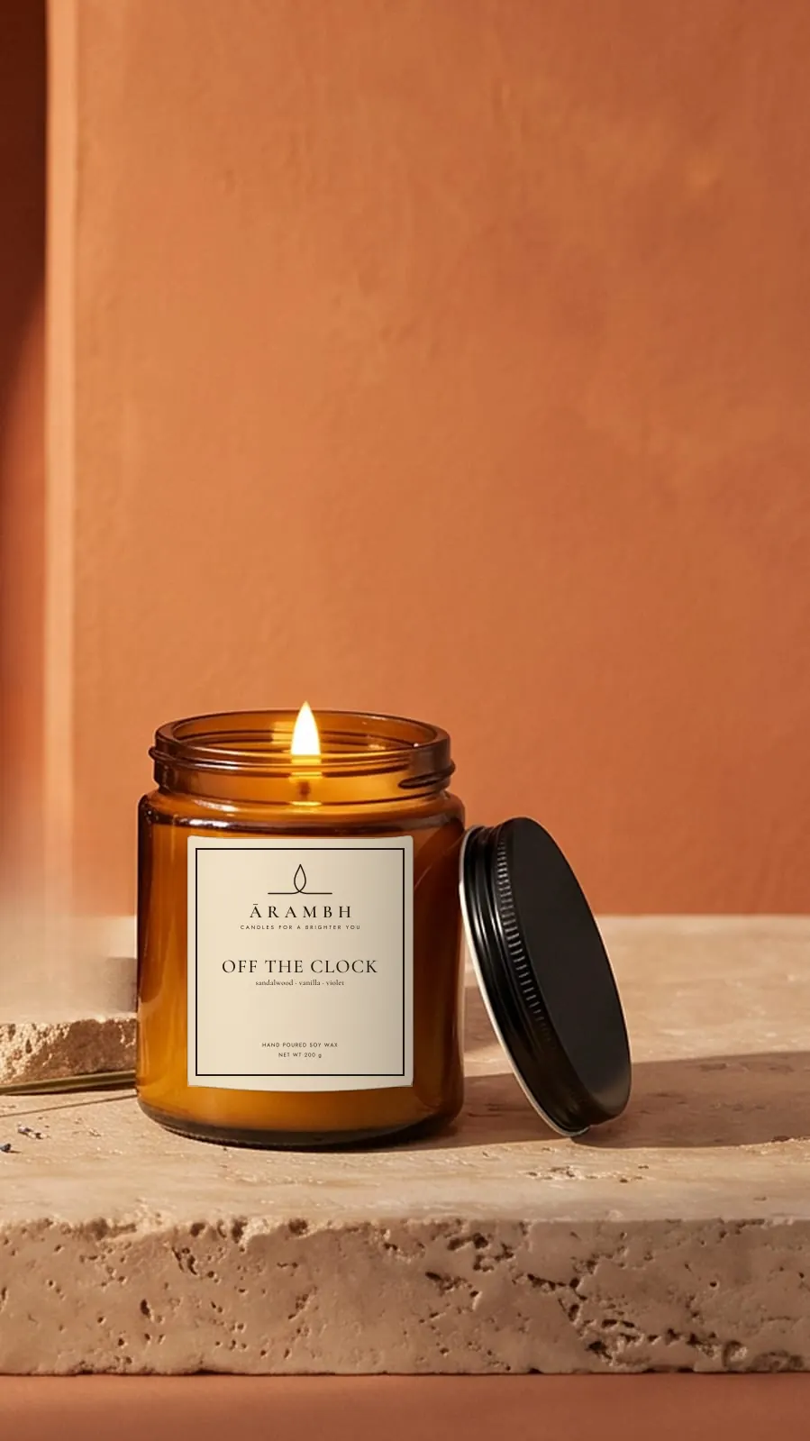A lit Off the Clock candle in an amber glass jar with the ĀRAMBH label, its black lid resting beside it on travertine
